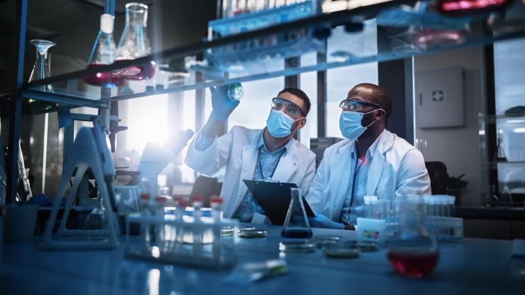 Two Scientists Wearing Face Masks use Microscope, Analyse Sample in Petri Dish, Discuss Innovative Technology